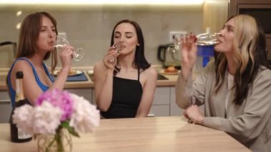 Üç kız modern bir mutfakta masada otururken uzun bir sapta uzun ince bardaklardan beyaz köpüklü şarap içerler. Kızlar konuşup gülümsüyor.