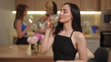 Ön planda, güzel beyaz bir kız bir bardaktan beyaz köpüklü şarap içiyor, arkadaşlarının arka planına karşı modern bir dairede mutfakta konuşuyor.