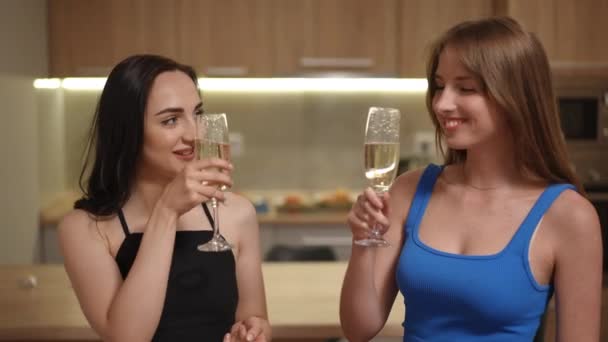 2人の女の子がアパートのモダンなキッチンに立っている間 白いスパークリングワインで眼鏡をかけます 女の子はメガネを飲んで お互いを見つめ合っている カメラを見て微笑んでいる若い女性 — ストック動画
