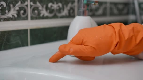 クローズアップ オレンジ色のゴム製グローブの手は 指で現代の家の浴室の白い洗面台の上のリムスケールの堆積物をこすろうとします — ストック動画