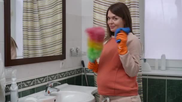 一个心情顽皮的孕妇看着摄像机 站在家里的浴室里 一个女人手里拿着吹笛器和一块破布 这个女人手上戴着橙色的橡胶手套 — 图库视频影像