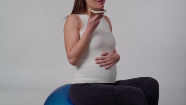 一个无法辨认的孕妇手里拿着一部手机 站在一个白色的背景上 坐在一个充气的体操球上 一边说着话 女人说话 抚摸她 — 图库视频影像