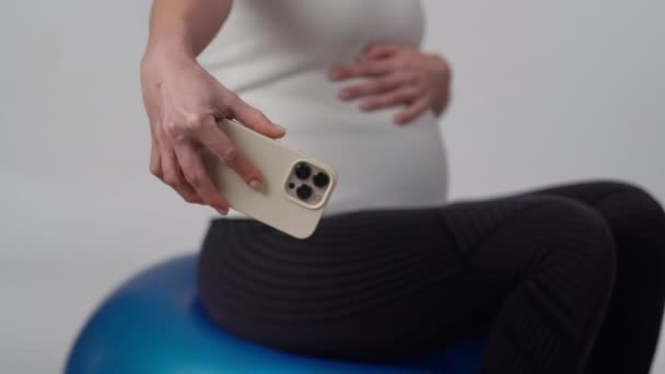 認識できない妊婦は携帯電話のフロントカメラで腹を撮り 彼女は白い背景に体操ボールの上に座っている間に腕の長さを保持します — ストック動画