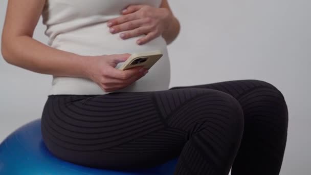 Nierozpoznawalna Ciężarna Kobieta Siedząca Piłce Gimnastycznej Białym Tle Trzyma Telefon — Wideo stockowe