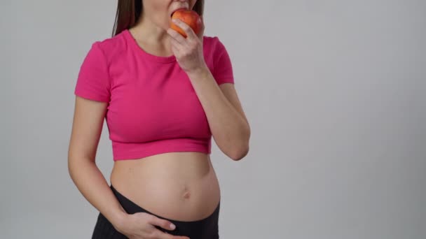 認識できない妊婦は白い背景に立って 手に持っているリンゴを食べます 女性は微笑む — ストック動画