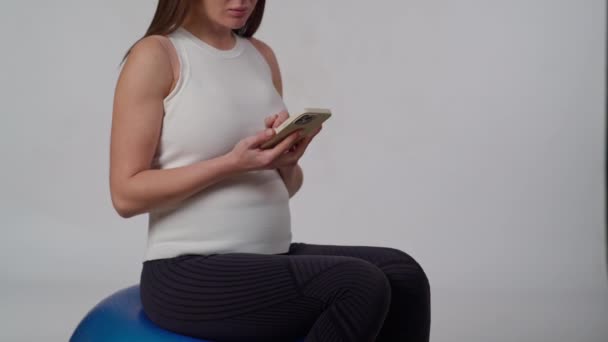 难以辨认的孕妇坐在一个白色背景的体操球上 手里拿着手机 手上拿着短信 面带微笑 — 图库视频影像