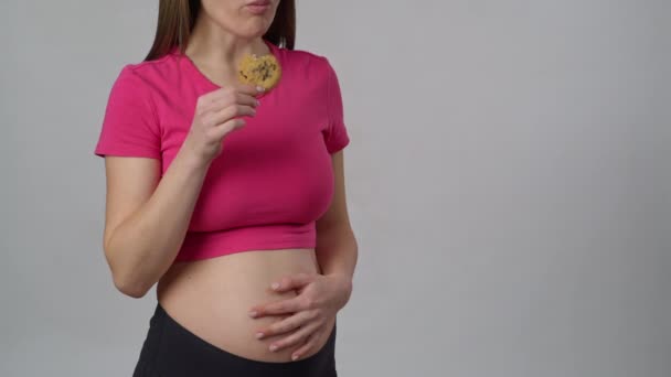 認識できない妊婦は白い背景に立って 手に持っているクッキーを食べます 女性は彼女の手と笑顔で彼女の腹を打つ — ストック動画