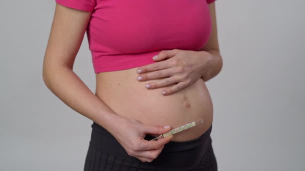 クローズアップ 認識できない妊婦は白い背景に立って 片手に水銀の温度計を保持し 他の人と穏やかに彼女の腹をストロークします — ストック動画