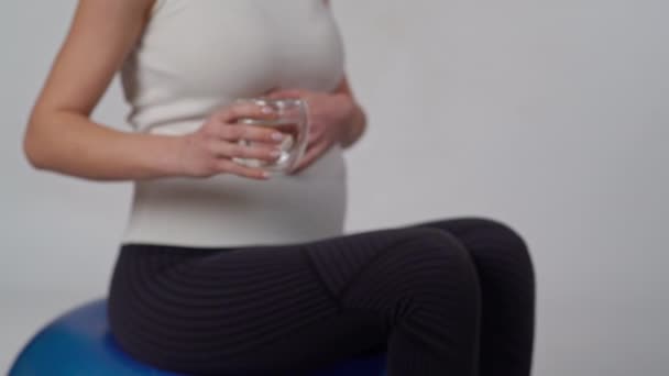 在摄像机前 一位孕妇手里拿着一杯水 坐在一个白色背景的充气体操球上 — 图库视频影像
