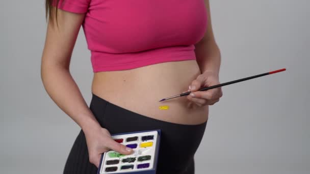 一个无法辨认的孕妇站在白色的背景上 手里拿着水彩画 用刷子在肚子上画着蓝色和黄色的条纹 — 图库视频影像