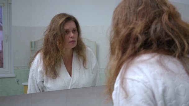白いバスローブの女性は トイレの壁の鏡で彼女の顔を注意深く見ています 肩の後ろから撮影 — ストック動画