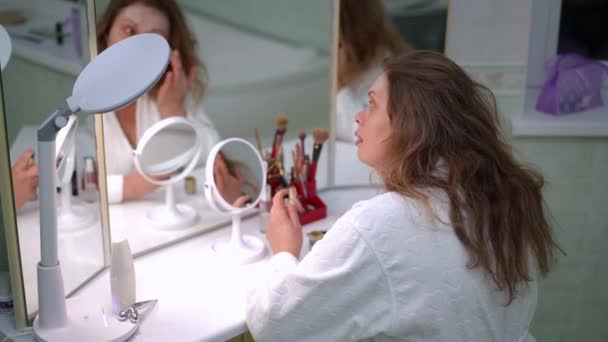 Женщина Наносит Крем Лицо Пальцем Сидя Белом Халате Перед Зеркалом — стоковое видео