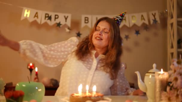 慢动作一个头戴生日帽的女人坐在节日桌旁跳舞 生日蛋糕上的蜡烛都烧焦了 今天是女人的生日 她既快乐又微笑 — 图库视频影像