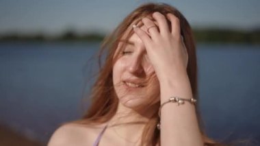 Yakın plan. Kız, parlak güneşten gözlerini kapayarak, uzun saçlarını eliyle yüzünden temizler. Nehir kıyısında dururken gülümseyen bir kız kameraya bakar.