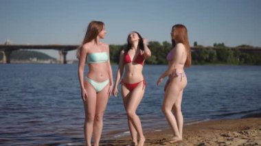 Ağır çekim. Mayolu üç kız nehrin kumlu kıyısında poz veriyor. Kızlar ellerini kaldırır, zıplar ve kameraya gülümser.