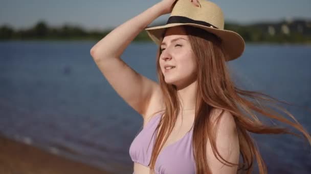 ピンク水着の女の子は 彼女の頭にレンガで軽い帽子を置き 強い風のために 川岸に立っている彼女の手でそれを保持します — ストック動画