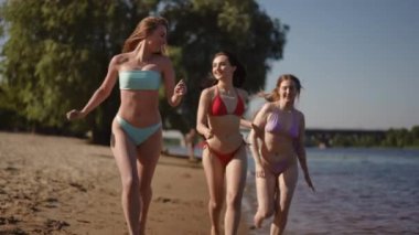 Ağır çekim. Üç bikinili kız nehrin kumlu kıyısında kameraya doğru koşuyor ve gülümsüyor.