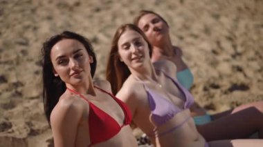 Uzun saçlı, mayo giymiş üç kız nehir kenarında kumlu bir sahilde oturuyorlar ve güneşleniyorlar.