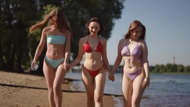 Három Bikinis Lány Sétál Homokos Folyóparton Kéz Kézben Mosolyog — Stock videók