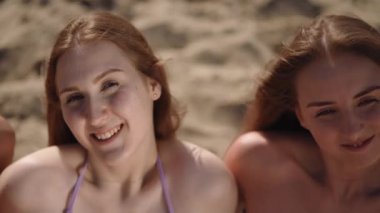 Yakın plan. Uzun saçlı, mayo giymiş üç kız sıcak, yaz, güneşli bir günde kumlu bir sahilde oturur ve güneşlenirler.