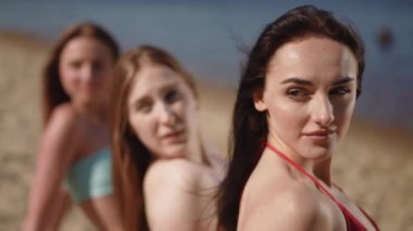 Yakın plan. Üç mayo giymiş kız, kumlu bir sahilde, güneşlenen bir battaniyede oturuyor. Kızlar omuzlarının üzerinden kameraya bakıyor.