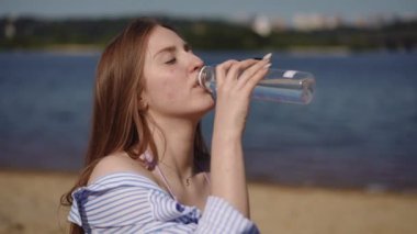 Bir kız nehir kıyısındaki kumlu bir sahilde otururken plastik bir şişeden su içiyor.