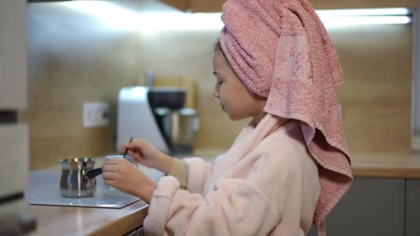 一个穿着浴衣 头上披着粉红毛巾的女孩 把一茶匙的碎咖啡洒在电炉上的一个煤气炉里 一个孩子站在屋里时在炉子上转动着 — 图库视频影像