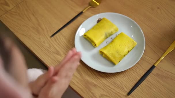 トップビュー バスローブにいる子供はテーブルに座り そこには2つのパンケーキが入ったプレートがあります 少女はフォークとナイフを拾い 朝食を食べようとしている — ストック動画