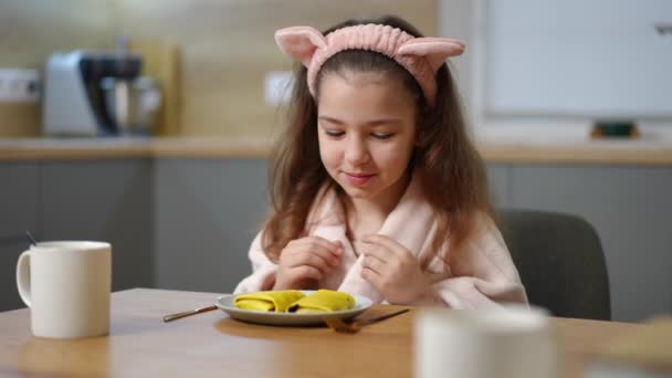 スローモーション バスローブの女の子は おいしい朝食を期待して手をこすり 2つの満たされたパンケーキのプレートがあるテーブルに座ります 少女はフォークとナイフを拾う — ストック動画