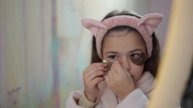 Yakın plan. Kafasında kedi kulağı olan pembe bir makyaj bandı olan bir kız aynanın karşısına oturur ve gözlerinin altına yamalar yapıştırır.