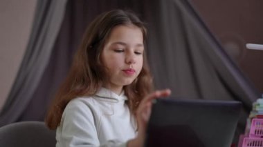 Küçük bir kız masanın üzerinde duran tablete bakıyor, gülümsüyor, elini sallıyor ve konuşuyor. Bir çocuk yatak odasında otururken tablet kullanarak iletişim kurar.