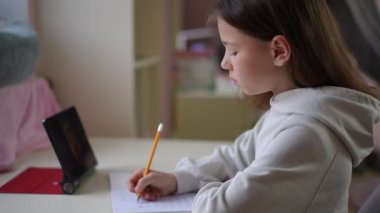 Modern online öğrenme. Küçük bir kız masaya oturur ve bir egzersiz kitabına kalemle yazar. Üzerinde online ders olan bir tablet masanın üzerinde duruyor. Çocuk tablet ekranına bakar