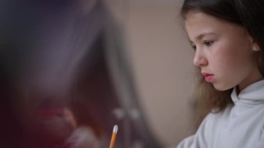 Dikkatlice bakıp kalemle yazan bir kızın yüzünün profili. Modern online öğrenme. Bir çocuk tablet ekranına bakar ve evde çevrimiçi öğrenme görevini tamamlar