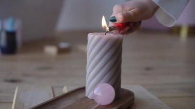 Yakın plan. Çakmak taşıyan bir kadın eli fitili büyük şekilli bir mumla yakar, ahşap bir standın üzerinde duran, kapalı alanda pembe bir kuvars topuyla.