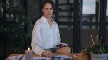 Dişi bir astrolog masadan bir tarot kartı alır ve ofisinde masasında otururken arkasında ne yazdığını okur. Kadın kameraya bakarken gülümsüyor. Yakın plan.