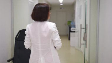 Beyaz önlüklü bir kadın hastane koridorunda dikilirken görülüyor. Beyaz ceketli bir kadın doktor dönüp dikkatlice bakar.