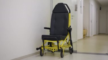 Parlak bir hastane koridorunda, cam kapıların yanında hasta nakil koltuğu var. Sandalye hastaları dışarıya ve ambulansın içine taşımak için maksimum güvenlik ve konfor sunuyor.