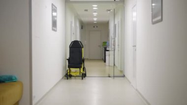 Kamera yavaşça sarı bir kanepe ve hasta nakil koltuğuyla aydınlık bir hastane koridoruna bakıyor.