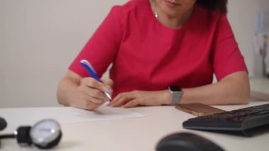 Yakın plan. Bir kadın masada oturuyor, bir kağıt parçasına yazarken konsantre oluyor. Kamera onun elini kağıt üzerinde hareket ederken gösteriyor, yazma eylemini yakalıyor..