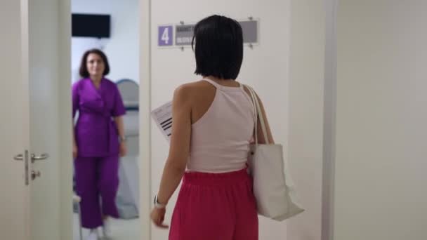 一个女人拿着一张纸走近妇科医生的办公室 在那里一位女医生正在敞开着的门口等她 女人进门时互相问候 随手关上门 — 图库视频影像