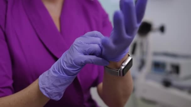 クローズアップ 紫色の医療スーツの認識できない女性医師は 医療機器付きの部屋に座っている間 紫色のゴム手袋に置きます — ストック動画
