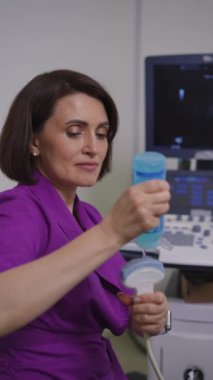Dikey video. Mor takım elbiseli kadın bir doktor ultrason taramasının önünde mavi plastik bir şişeden ultrason sondasına şeffaf bir jel uyguluyor.