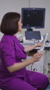 Dikey video. Mor takım elbiseli kadın bir jinekolog ultrason makinesinin yanında oturur ve elinde hazır bir ultrason sensörü tutar, kameraya bakar ve gülümser. Ultrason: