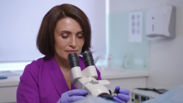クローズアップ 紫色の医療用スーツの女性婦人科医が眼鏡の上に曲がり 双眼鏡のコルポスコープを通して見ています 現代のクリニックでのコルポスコピー手順 — ストック動画