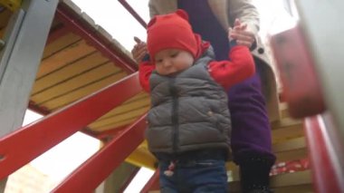 Tanınmayan bir kadın, küçük bir çocuğu tırmanma merdivenlerinden indirip kollarından tutuyor. Anne ve oğlu bir bahar günü şehir parkında oynuyorlar.