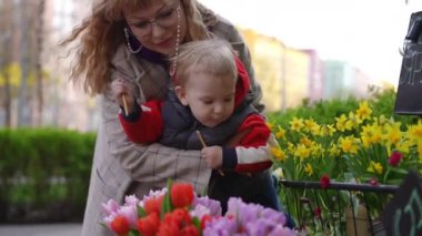 Ağır çekim. Bir anne, kucağında küçük oğluyla bir bahar günü şehirdeki bir çiçek fuarında dikilirken çiçekleri koklamak için eğilir. Küçük bir çocuk tuzlu çubuk yiyor, elinde tutuyor.