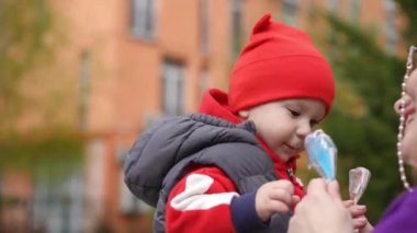 Ağır çekim. Yakın plan. Anne küçük çocuğa elinde iki plastik oyuncak balık veriyor ve o da dikkatlice alıyor. Anne ve oğlu bir bahar günü arka planda bir binayla birlikte dışarıdalar.