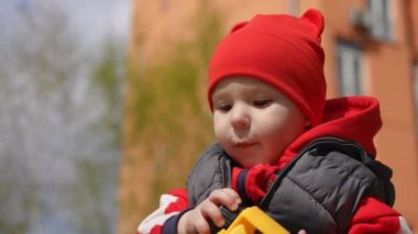 Ağır çekim. Yakın plan. Kırmızı çocuk beresi takmış küçük bir çocuk güneşli bir bahar gününde şehir parkında plastik oyuncak kazıcısıyla oynuyor.
