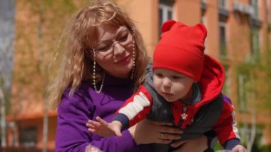 Ağır çekim. Yakın plan. Bir anne, oyuncak bir balığa uzanan küçük oğlunu kollarında tutar. Anne ve bebek güneşli bir bahar gününde bir yerleşim yerinin avlusunda