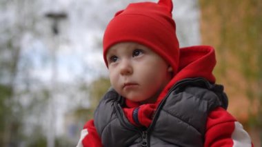 Ağır çekim. Yakın plan. Bir bahar günü şehir parkında açık havada oturan, kırmızı bereli, düşünceli, küçük bir çocuğun bakışları.